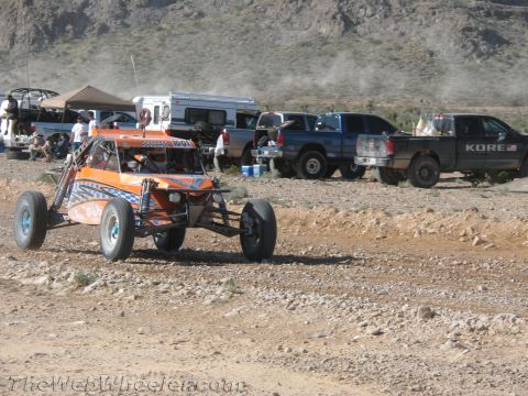 1990 Ford Bronco - orange open-frame off-road buggy numbered 1001 drives a dirt course as pickups and a canopy line the pit behind it.