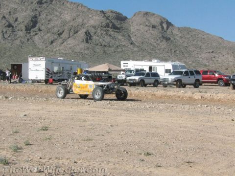 1990 Ford Bronco - open-frame off-road buggy numbered 1001 sits in a desert pit area backed by RVs, enclosed trailers and parked SUVs below rocky hills.