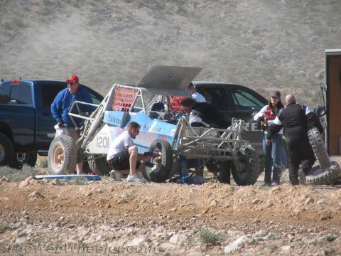 1990 Ford Bronco - crew works on a blue open-frame off-road buggy numbered 1201 in a dusty desert pit, with a pickup and tow rig parked nearby.
