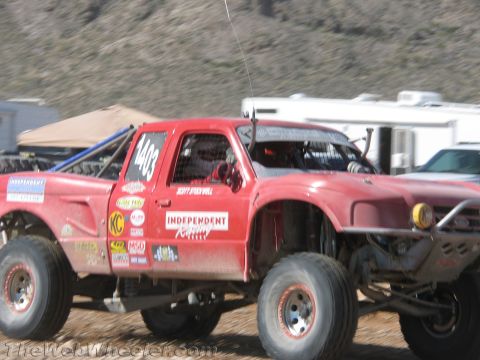 1990 Ford Bronco - red off-road race truck numbered 403 and lettered 'Independent Racing' drives a dirt course with trailers and hills behind.
