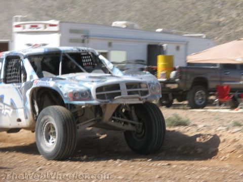 1990 Ford Bronco - silver-and-blue off-road race truck corners on a dirt track, motion-blurred, with a white enclosed trailer and SUV in the background.