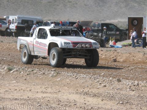1990 Ford Bronco - white off-road race truck numbered 703 drives a dirt course as spectators, pickups and a KORE truck sit along the pit behind.