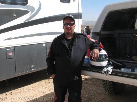 1990 Ford Bronco - man in a black racing suit, cap and sunglasses leans on a pickup tailgate holding a helmet and gloves, an RV at his side.
