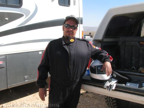 1990 Ford Bronco - smiling man in a black racing suit, cap and sunglasses leans on a pickup tailgate with a helmet and gloves beside an RV in the desert.