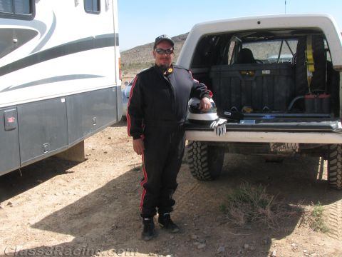 1990 Ford Bronco - man in a black-and-red racing suit, cap and sunglasses stands beside an open pickup tailgate, with an RV and desert hills behind.