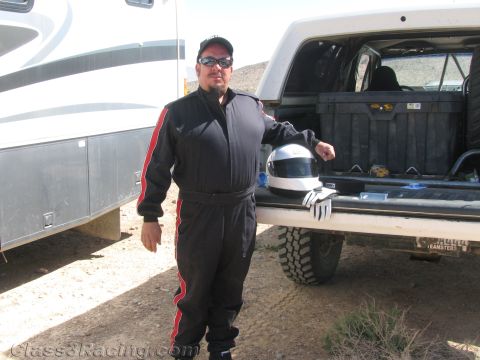 1990 Ford Bronco - man in a black-and-red racing suit, cap and sunglasses leans on a pickup tailgate with a helmet and gloves, an RV alongside.