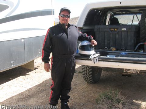 1990 Ford Bronco - smiling man in a black-and-red racing suit, cap and sunglasses stands by a pickup tailgate holding a helmet and gloves beside an RV.