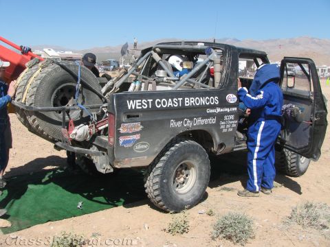1990 Ford Bronco - black tube-chassis off-road race Bronco lettered "West Coast Broncos" sits in a desert pit while a crew member in blue works at the open do…