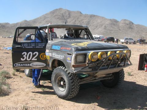 1990 Ford Bronco - weathered off-road race truck numbered 302 with a light bar and Deaver Suspension lettering sits in a dusty desert pit near mountains.