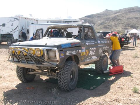 1990 Ford Bronco - Off-road race truck number 302 sits on a green tarp in a desert pit as a crew member in yellow works at the rear, with RVs behind.