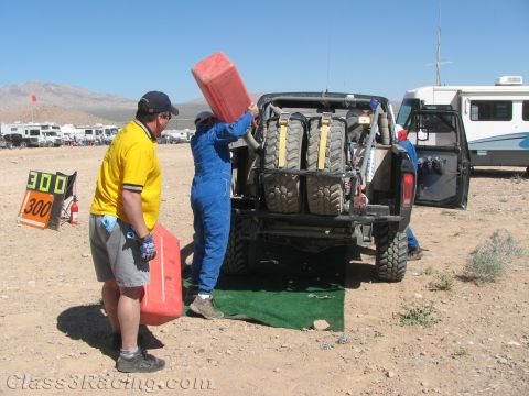 1990 Ford Bronco - man in blue overalls pours fuel from a red jug into a rear-engine off-road race vehicle while a helper in yellow waits with another jug.