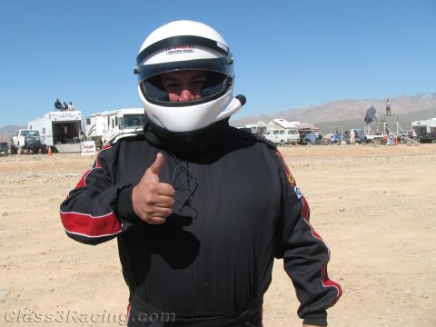 1990 Ford Bronco - race driver in a black firesuit and white full-face helmet gives a thumbs-up in a desert pit area under clear sky.