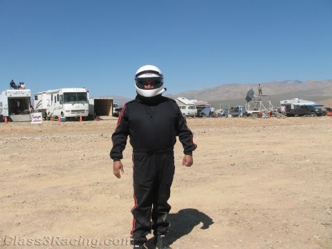 1990 Ford Bronco - race driver in a black firesuit and white helmet stands in a dusty desert pit with RVs and mountains in the background.