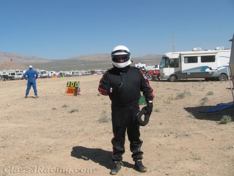 1990 Ford Bronco - race driver in a black firesuit and white helmet holds his gloves while standing near a 300 course marker in the desert.