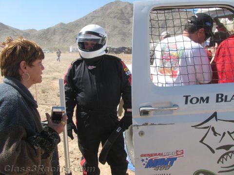 1990 Ford Bronco - driver in a black firesuit and helmet talks with a woman beside a white off-road race truck lettered "Tom Blair" in the desert.