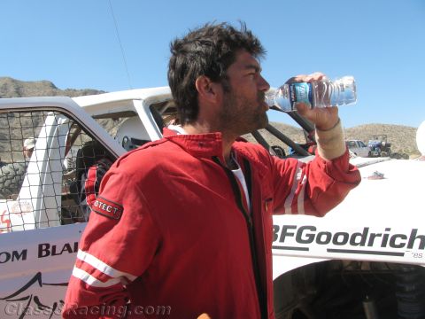 1990 Ford Bronco - man in a red racing jacket drinks from a water bottle beside a white off-road race truck with BFGoodrich lettering in the desert.