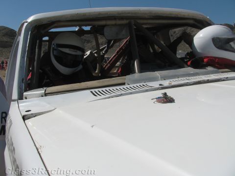 1990 Ford Bronco - Looking over the white hood of a race truck through the windshield reveals the roll cage interior and seated occupants in white and red hel…