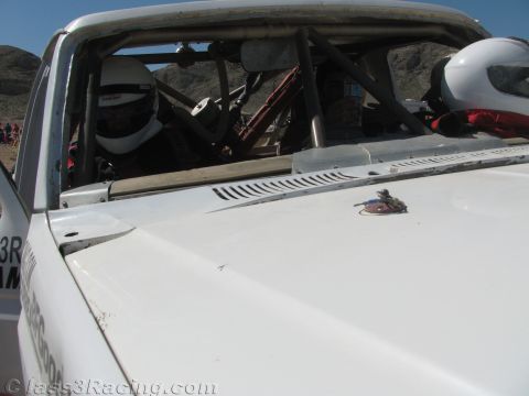 1990 Ford Bronco - view over the white hood of an off-road race truck into the roll-caged cabin where two occupants wear white and red helmets.