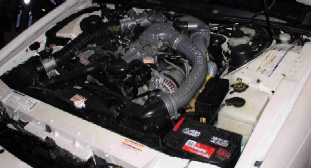 White car's engine bay with supercharged V6, black intake tubing and battery