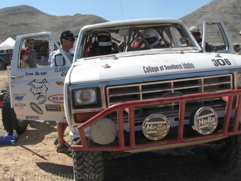 1990 Ford Bronco - white Ford off-road race truck numbered 306 lettered "College of Southern Idaho" with a red brush guard and Hella lights sits in a desert p…