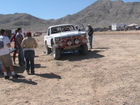 1990 Ford Bronco - white off-road race truck rolls across an open desert course as a group of spectators watches near rugged mountains.