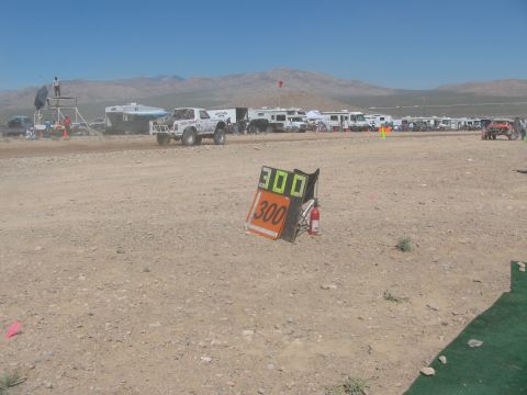 1990 Ford Bronco - orange and green "300" course marker board with a fire extinguisher stands in the desert with race vehicles and RVs in the distance.