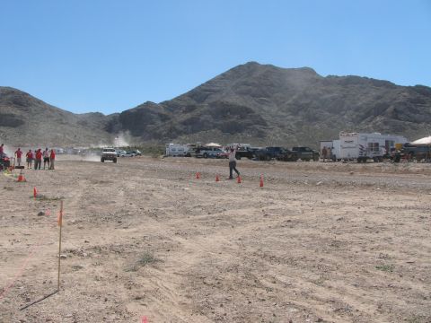 1990 Ford Bronco - coned desert race course stretches toward a mountain as spectators in red watch and dust rises from a distant vehicle.