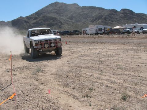 1990 Ford Bronco - white off-road race truck kicks up dust as it drives a coned desert course with RVs and a mountain behind.