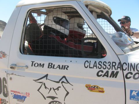 1990 Ford Bronco - Two helmeted occupants sit behind the window mesh of a white off-road race truck lettered "Tom Blair" and "Class3Racing" with a dog-face lo…