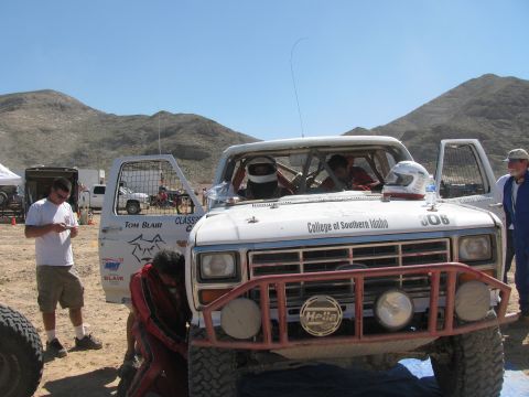 1990 Ford Bronco - front of white Ford race truck 306 "College of Southern Idaho" with helmeted occupants while crew and spectators gather in the desert.