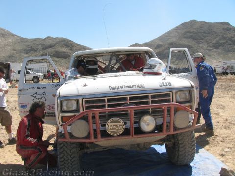 1990 Ford Bronco - White Ford race truck 306 "College of Southern Idaho" sits with doors open as crew in red firesuits work around it in a desert pit.
