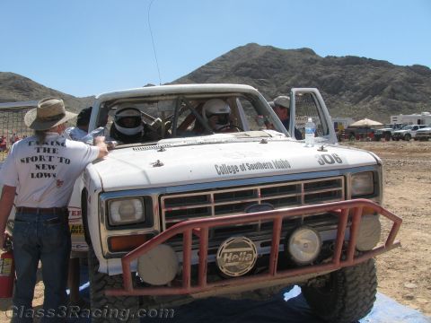 1990 Ford Bronco - White Ford race truck 306 in a desert pit as a man in a hat wears a T-shirt reading "Taking the Sport to New Lows."