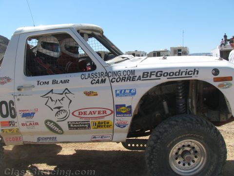 1990 Ford Bronco - Side view of a white off-road race truck lettered "Tom Blair," "Cam Correa" and "BFGoodrich" with helmeted occupants in a desert pit.
