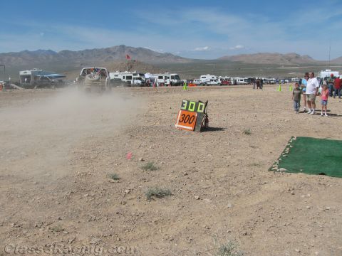 1990 Ford Bronco - 300 course marker board stands on a desert course as a vehicle raises a dust trail and spectators including children watch nearby.