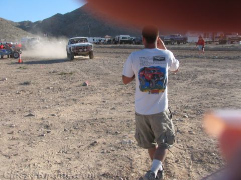 1990 Ford Bronco - man in a graphic T-shirt photographs a white off-road race truck raising dust on a desert course in warm late-day light.