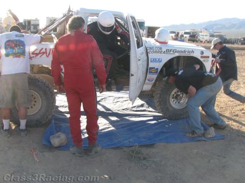 1990 Ford Bronco - pit crew, one in a red driving suit and another in a helmet, tends to a white off-road race truck on a tarp in the desert.