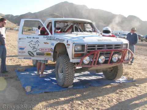 1990 Ford Bronco - white desert race truck with a red tube bumper, round driving lights and sponsor decals sits in a pit area with crew nearby.