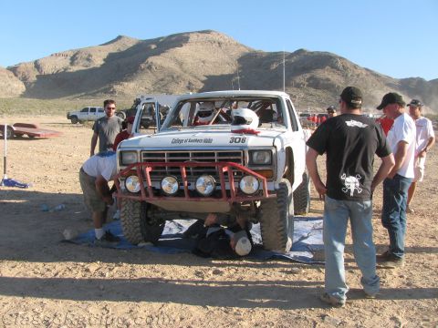 1990 Ford Bronco - white desert race truck numbered 306 with a red tube bumper and round lights is surrounded by people in a dusty pit.