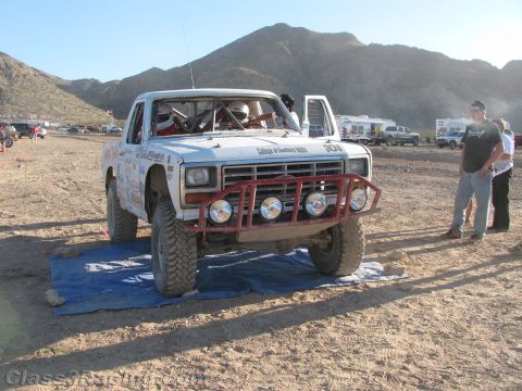 1990 Ford Bronco - white desert race truck numbered 306 with a red tube bumper sits in a pit area as two people stand alongside.