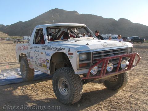 1990 Ford Bronco - white off-road race truck with a red tube bumper, round lights and visible roll cage waits in a desert pit with spectators around.