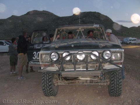 1990 Ford Bronco - Head-on dusk view of a dark off-road race truck numbered 302 with a roof light bar and tube bumper, people gathered around.