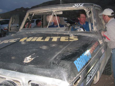 1990 Ford Bronco - Close view of a dirty dark race truck hood marked KC HiLiTES and Bronco, with the driver and others visible through the windows.