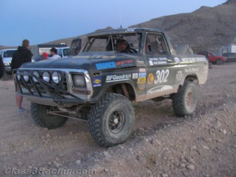 1990 Ford Bronco - black desert race truck numbered 302 with round front lights and a tube bumper sits on rocky ground at dusk.