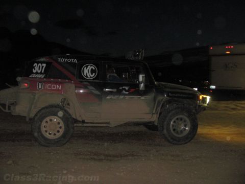 1990 Ford Bronco - dark off-road race truck numbered 307 with Toyota and ICON decals drives at night near a parked trailer.