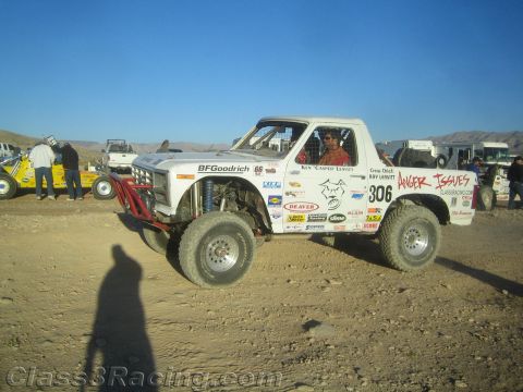1990 Ford Bronco - white desert race truck numbered 306 lettered Anger Issues with a driver seated, parked on dirt with spectators behind.