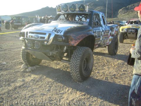 1990 Ford Bronco - Ford off-road race truck numbered 1401 with flame graphics, a roof light bar and tube bumper in a busy desert staging area.