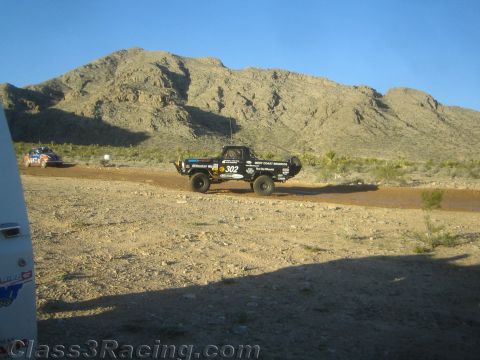 1990 Ford Bronco - dark off-road race truck numbered 302 drives along a desert dirt road with rocky mountains rising behind.