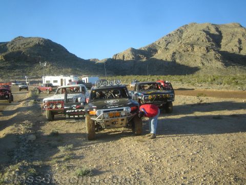1990 Ford Bronco - Several off-road race trucks, including a black one with KC lights, line up in a desert staging area beneath rocky mountains.