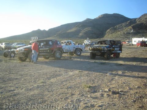 1990 Ford Bronco - row of off-road race trucks, including a white number 306 and a blue number 302, parked on desert dirt with crews nearby.