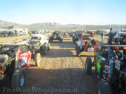 1990 Ford Bronco - crowded field of off-road buggies and trucks, including numbers 1302 and 1305, lines a gravel staging corridor in the desert.
